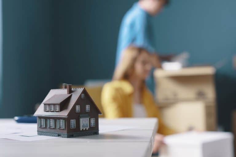 A house figurine and a young couple and boxes in the background in the concept of packing for a house move.