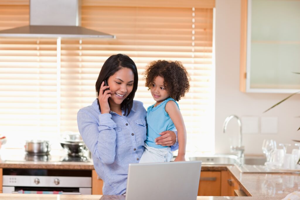 A mother is using her phone while in front of the laptop while carrying her daughter