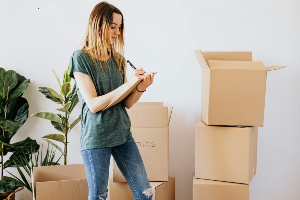 A woman is writing on a clipboard while being surrounded by cardboard boxes