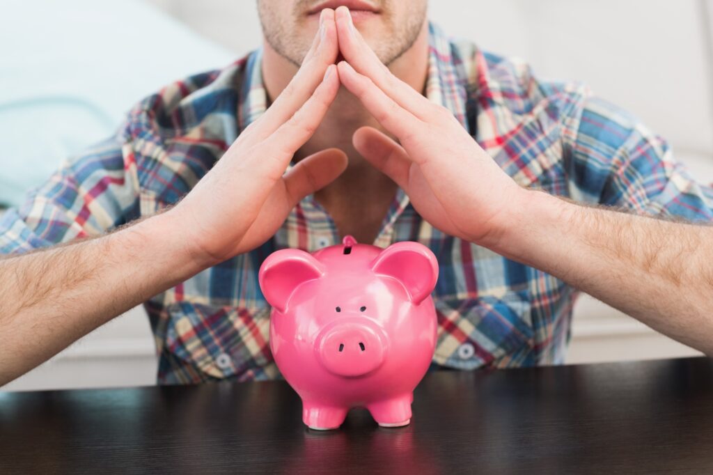 A man with his hands forming a triangular roof of a house and a piggy bank in the concept of 'how to save money when moving to Twickenham'.
